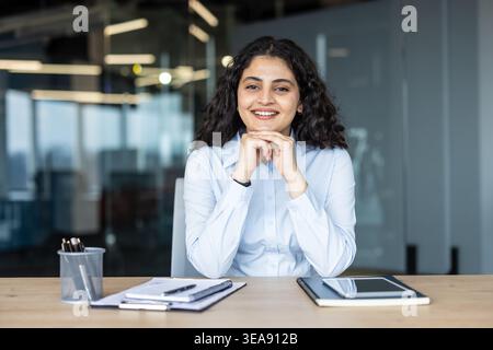 Young businesswoman with dark curly hair smiling confidently, sitting at a modern office desk with paperwork and a tablet, representing professional success and leadership Stock Photo