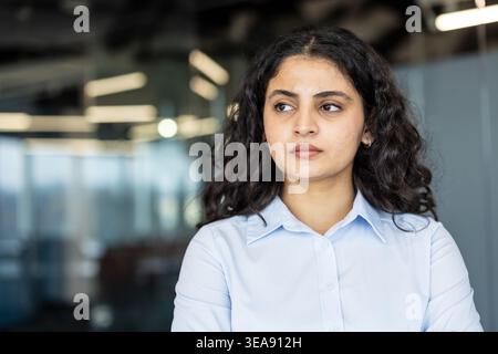 Thoughtful indian businesswoman with curly dark hair in a modern office, standing in smart casual attire, looking away as she contemplates strategy, ideas and future plans Stock Photo