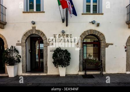 Entrance to Hotel Centrale in Olbia, Sardinia, Italy, featuring stone-arched doorways, white facade, potted plants, and Italian and regional flags. Stock Photo