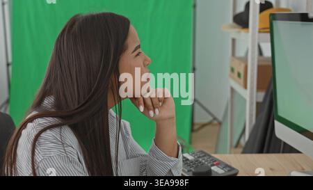 Young woman resting chin on hand beside dslr camera, keyboard and monitor in green screen studio; thoughtful focus. Stock Photo