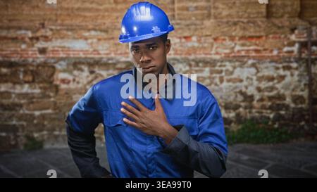 Man construction worker, african american, in blue hardhat and blue coveralls, places hand on chest in front of brick building, serious expression and Stock Photo