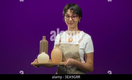 Woman wearing apron holds wooden tray of three ceramic vases, smiling and presenting them with both hands in studio; creative craft joy. Stock Photo