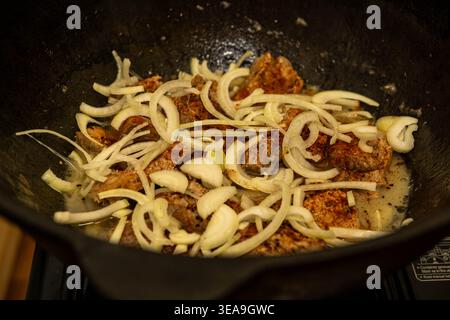 Close-up of seasoned meat chunks cooking with freshly sliced onions in a cast iron pot (kazan) on a portable stove. The dish is simmering. Stock Photo