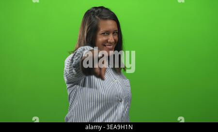 Woman smiling and points finger toward camera while standing against green screen in studio wearing a striped shirt; confidence. Stock Photo