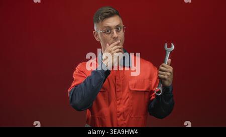Young mechanic man in orange coverall holds a wrench and places hand to mouth gesture, wearing glasses against a red studio backdrop while inspecting Stock Photo