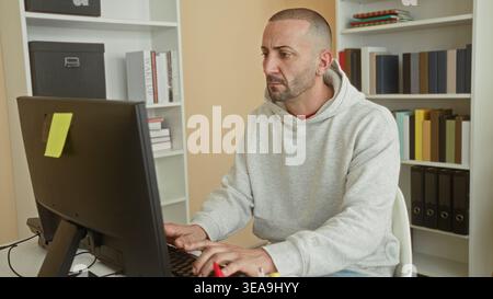 Man typing on computer, hands on keyboard, wearing hoodie at desk with monitor and bookshelves in building; focused productivity. Stock Photo