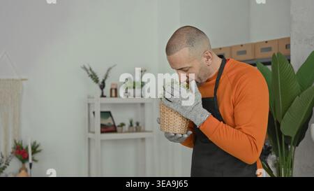 Man florist smelling a potted plant, nose close to tiny white flowers, holding wicker basket in a building; gentle care serenity. Stock Photo