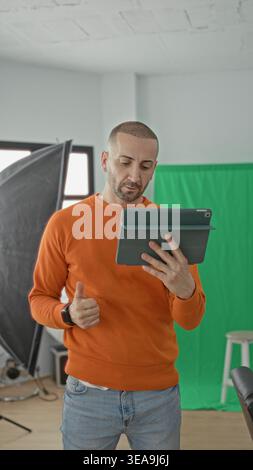 Man holding tablet and gesturing with hand in studio among softbox lights and green screen, wearing orange sweater and jeans; creative concentration. Stock Photo