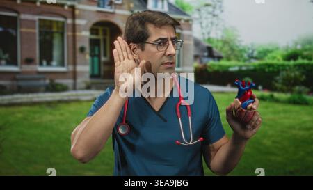 Man doctor in blue scrubs holds anatomical heart model and raises hand to ear in front of building; health awareness concern. Stock Photo