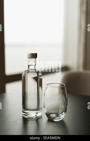 Clear glass bottle with a silver cap sits on a dark wooden table. An empty drinking glass is beside it. Soft natural light streams through a large win Stock Photo