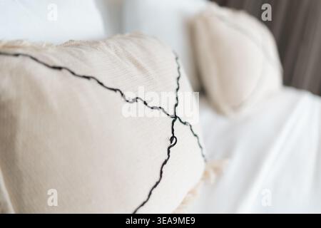 Close-up of beige pillow with black, wavy stitching. Soft white fabric forms the background. Pillow sits on a bed or sofa. Gentle texture adds comfort Stock Photo
