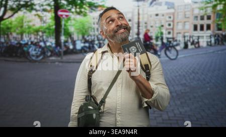 Man holding passport and gripping backpack strap on city street, smiling and looking upward; travel joy. Stock Photo