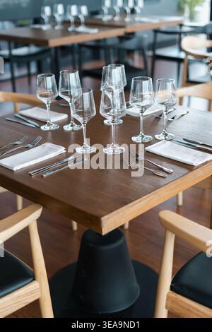 Wooden table holds empty wine glasses and silverware. White napkins rest beside polished cutlery. Light wood chairs surround the table. Setting implie Stock Photo