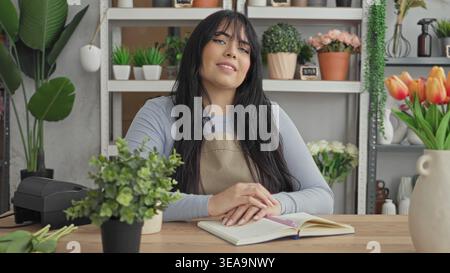 Woman florist in apron smiling with hands clasped on open notebook at studio counter, surrounded by potted plants, vases, shelves and tulips; welcomin Stock Photo