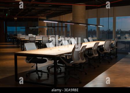 Modern office interior with long wooden tables and black metal frames. White ergonomic chairs surround each workspace. Large windows show city skyline Stock Photo