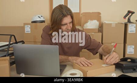 Man with blond hair writing a shipping label on a cardboard parcel with his hands at a cluttered desk with laptop, tape roll and stacked boxes in a pa Stock Photo