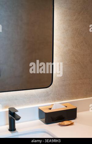 Black faucet gleams on the white sink basin. Dark tissue box rests on the countertop surface. Small soap dish holds a single bar of soap. Large mirror Stock Photo