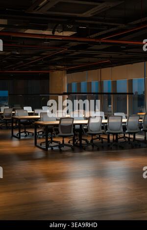Modern conference room has polished wooden floors. Long table with ergonomic office chairs around it. Exposed ceiling ducts and large windows fill the Stock Photo