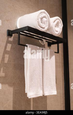 Black metal towel rack hangs on beige tiles. Two white towels roll on top. Two more hang below the rack. The setup looks tidy and modern. Suitable for Stock Photo