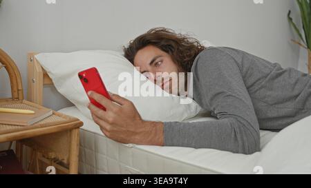 Man lying on bed holding red smartphone, hand and face visible, head on pillow in building; tired scrolling solitude. Stock Photo