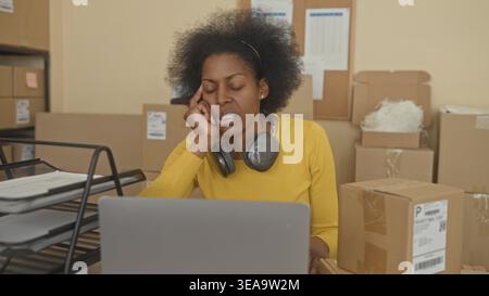 Woman with laptop, stacked parcels and headphones, hand on temple while sorting boxes at a packing building; stress overwhelm. Stock Photo