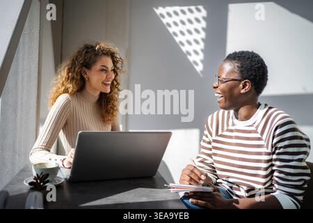 Diverse women friends collaborating and laughing in coffee shop Stock Photo