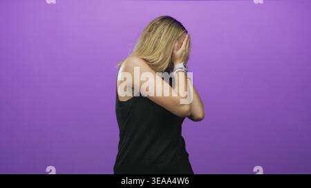 Woman shields face with outstretched hand and visible palm in studio with purple backdrop, wearing black tanktop and bracelet; shyness. Stock Photo