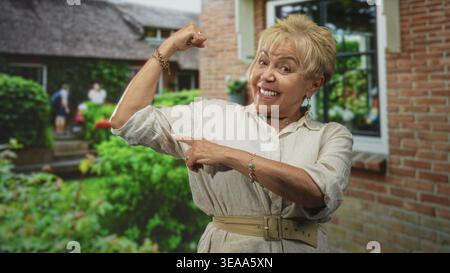 Senior woman flexes bicep and points to it outside a brick building doorway; confidence empowerment. Stock Photo
