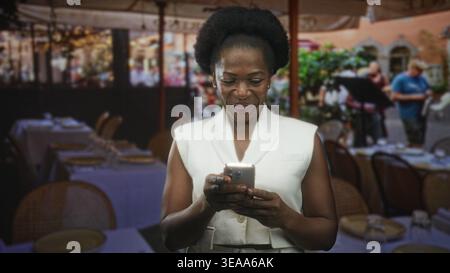 Woman holding smartphone, smiling and looking up among street cafe tables in a white vest and relaxed pose; calm joy. Stock Photo