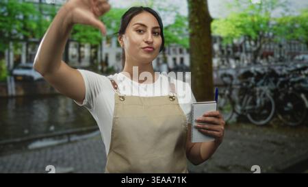 Woman waitress in apron holding notebook shows thumbs down on street in amsterdam; disapproval service feedback. Stock Photo
