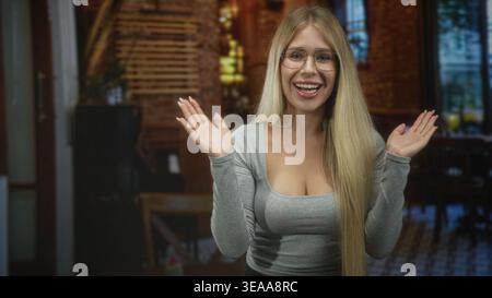 Young blonde woman with glasses smiling, hands covering mouth and gesturing, wearing grey fitted top, long hair, wooden staircase and tables in a cafe Stock Photo