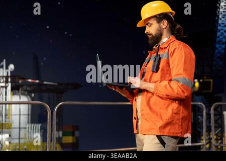 Offshore rig employee monitoring operations using laptop on oil platform deck late at night. Barge engineer wearing hardhat reviewing energy operations on drilling rig using notebook Stock Photo