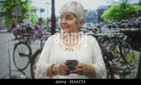Woman with grey hair holding smartphone in both hands on street, smiling while texting; contentment connection. Stock Photo