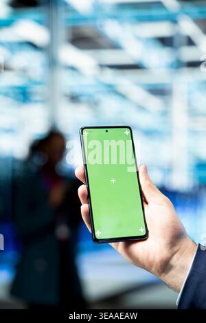 Configuring server settings, green screen smartphone closeup and engineer in data center. Using mockup mobile phone, infrastructure maintenance tasks and IT specialist in modern high tech server hub Stock Photo