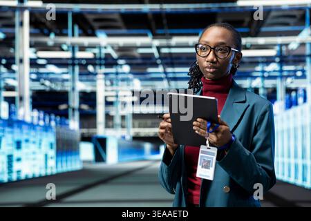 Data center female engineer uses tablet to do maintenance tasks. African american woman working in server room troubleshooting technical issues with device to ensure network uptime Stock Photo