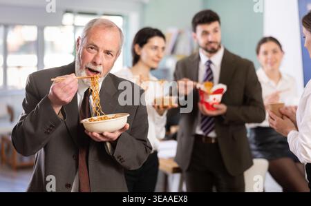 Mature businessman having takeout meal during lunch break at work with other colleagues in office Stock Photo