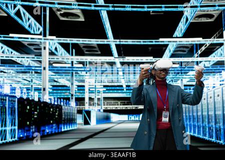Data center engineer uses VR headset tech to upgrade database storage equipment. African american woman uses virtual reality simulation in server hub to update cloud computing rigs, technology concept Stock Photo
