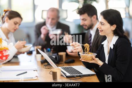 Lunch break - woman eating noodles with colleagues in modern office Stock Photo