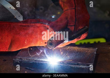welder wearing heavy-duty red leather protective gloves uses an electrode holder to perform stick welding on a thick metal plate, creating a bright ar Stock Photo