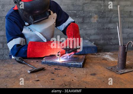 welder in full protective gear, including a mask, leather apron, and red gloves, uses a stick electrode to weld a steel plate in a workshop. Stock Photo