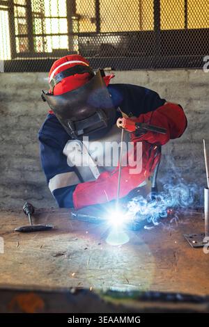 welder in full protective gear, including a mask, leather apron, and red gloves, uses a stick electrode to weld a steel plate in a workshop. Stock Photo