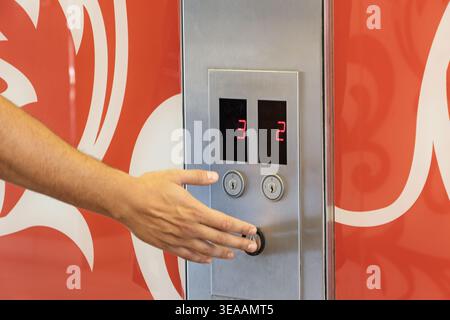 person reaches out to press the call button on a stainless steel elevator panel featuring two digital floor displays showing numbers two and three. Stock Photo