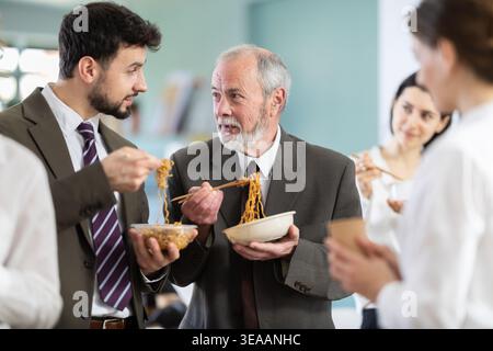 Positive two male office workers eating food together during lunch time in office of business company Stock Photo