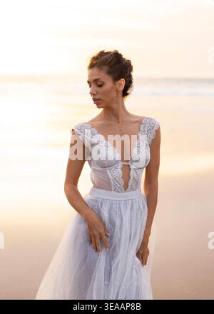 Young woman in white lace dress standing on beach at sunset with calm sea, showing elegance, natural beauty and peaceful lifestyle Stock Photo