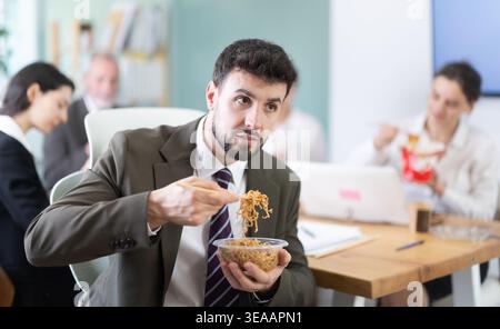Young businessman having takeout meal during lunch break at work sitting at workplace at laptop with other colleagues in office Stock Photo