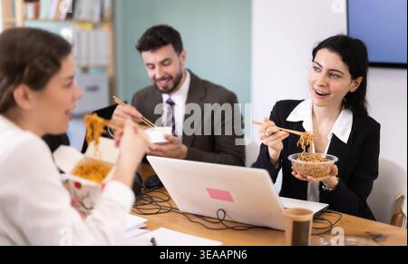 Office workers resting on break together eating, drinking coffee and talking, sitting at workplace at table with documents Stock Photo