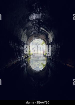 A dark stone brick arch tunnel on the Sandiaoling Three-Tiered Waterfall Trail in New Taipei, Taiwan, with a bright circular exit in the distance reflected in still water on the path. The moody leading lines and atmospheric perspective create a dramatic hiking scene. Stock Photo