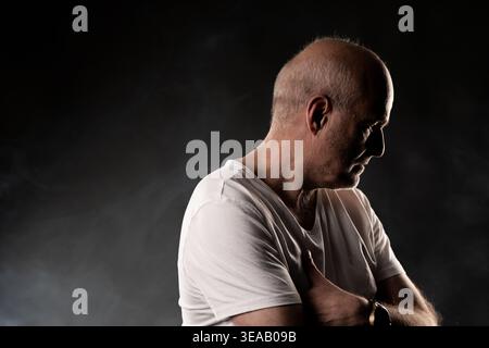 Mature bald man in profile looking down, contemplating in dark setting Stock Photo