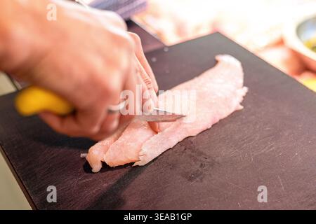 A hand is seen cutting a fillet of fish on a black cutting board in a kitchen. The person uses a knife. Slices of lemon are nearby to add flavor. This Stock Photo