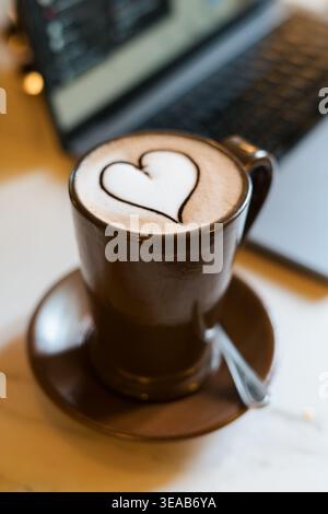 Dark ceramic coffee cup steams gently. White heart shape floats on the surface. Matching saucer holds the cup securely. Spoon rests neatly beside it Stock Photo